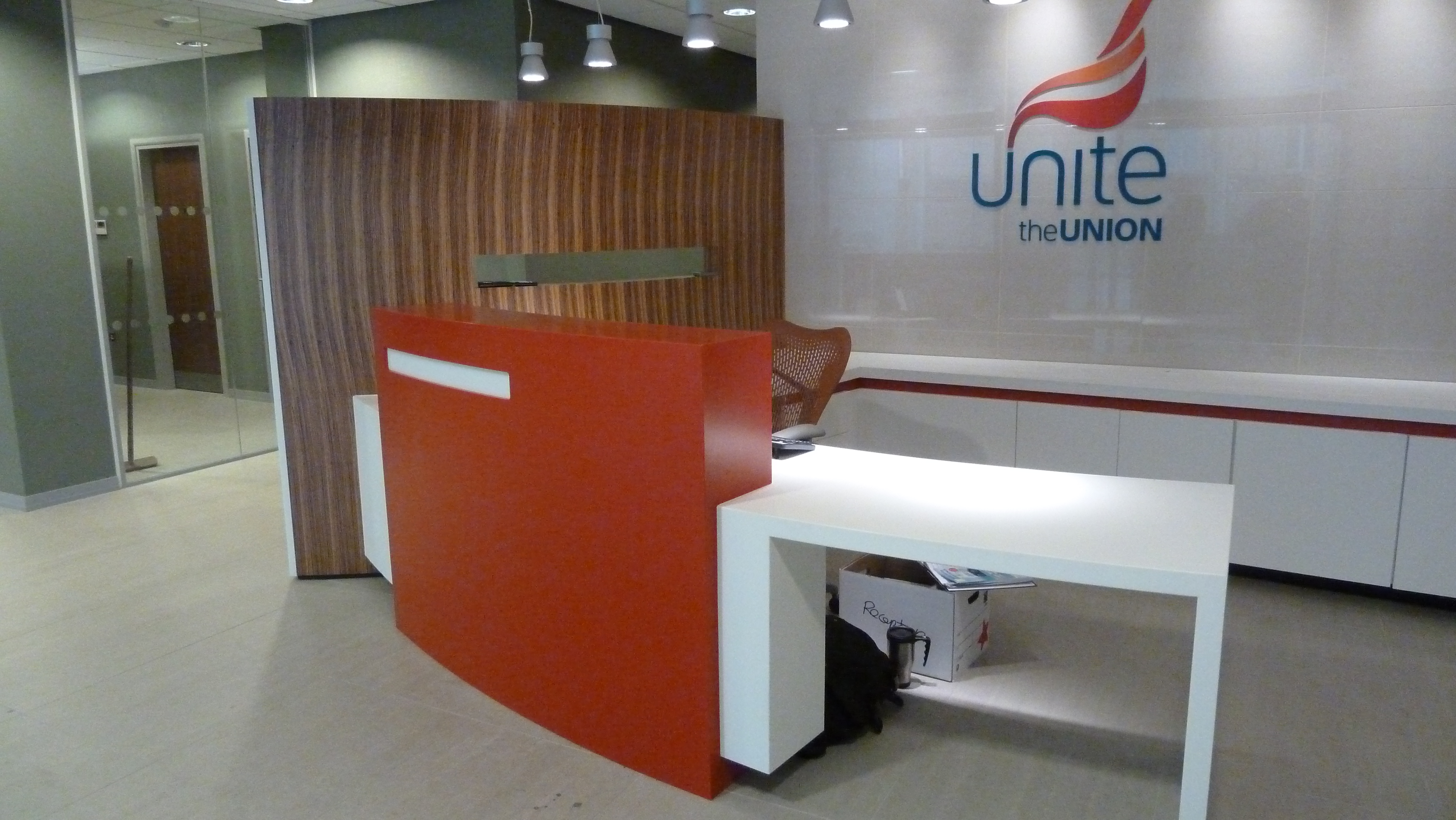 Unite the Union Reception Desk – Liverpool Manufactured from corian using hot and glacier white . Rear cupboards and worktops including doors manufactured from glacier white corian with hot (RED) inlay band. Curved laminated privacy screen wall made up from mdf and laminated in zebrano formica and a curved glass vision panel . 